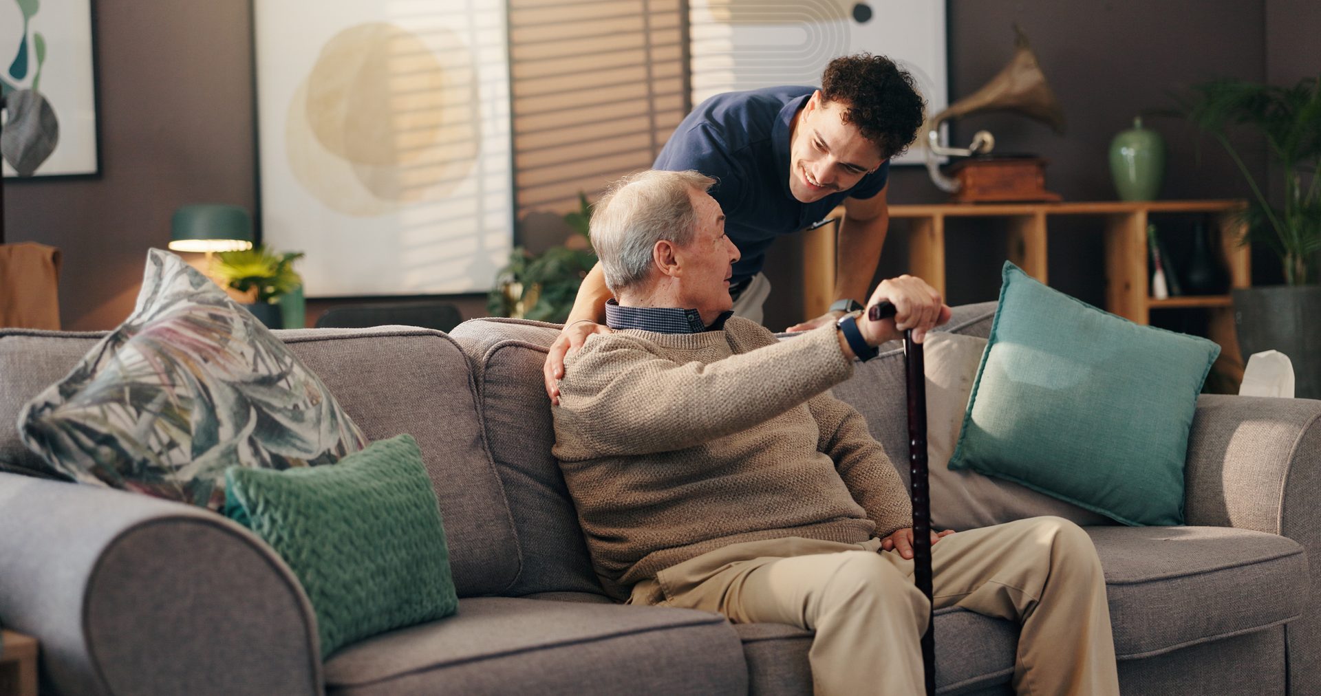 Caregiver helping a senior man with a cane rise from the sofa