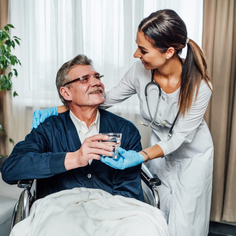 An older man in a wheelchair smiles at the nurse-assistant, she hands him a glass of water. Nursing home, help for the elderly.